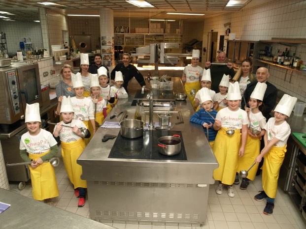 Kinder mit Kochmützen und Schürzen in der Küche mit Erwachsenen im Hintergrund